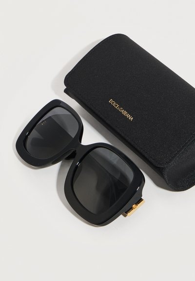 Μαύρα υπέρμετρα τετράγωνα γυαλιά ηλίου με σκούρους φακούς δίπλα σε μαύρη θήκη Dolce & Gabbana με χρυσά γράμματα πάνω σε λευκή επιφάνεια.