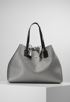 Grand sac à main argenté avec des poignées noires et un détail de nœud rayé noir et blanc sur un piédestal blanc sur un fond uni.