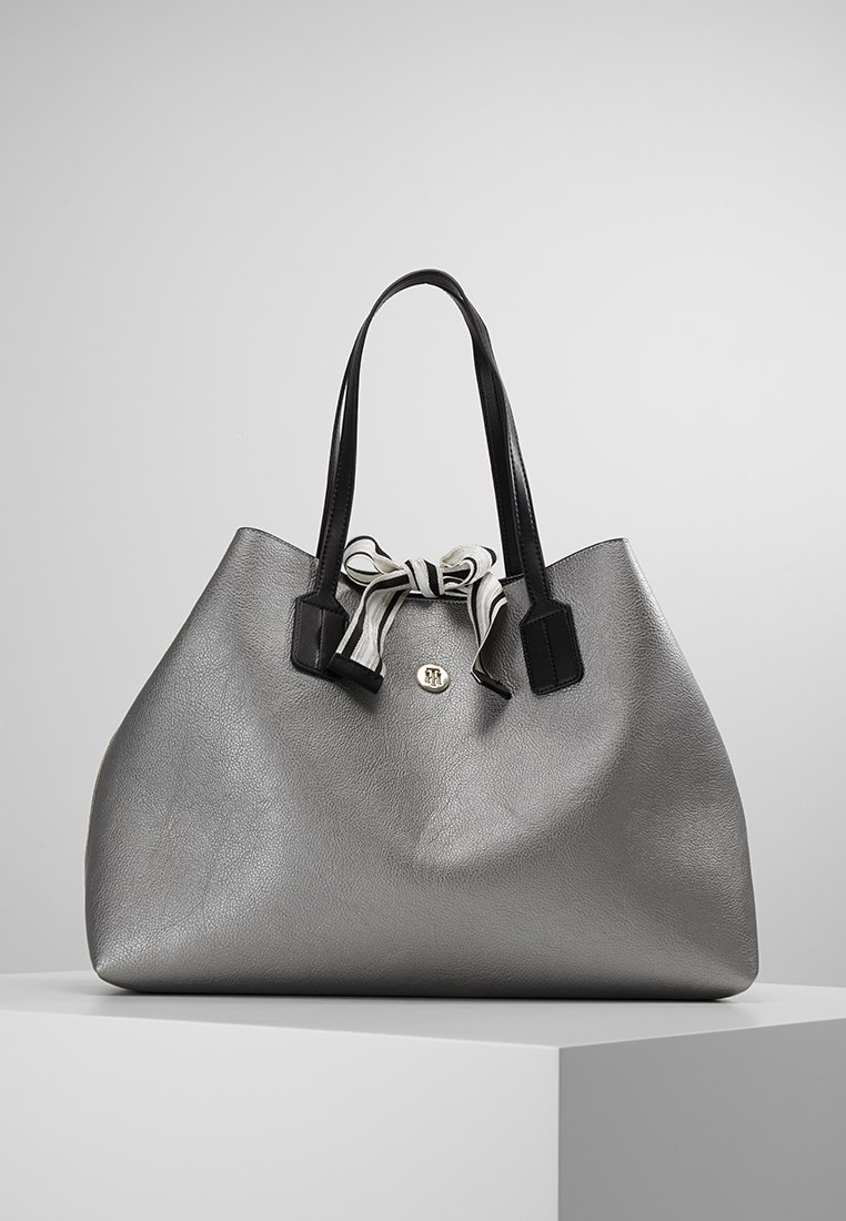 Grand sac à main argenté avec des poignées noires et un détail de nœud rayé noir et blanc sur un piédestal blanc sur un fond uni.