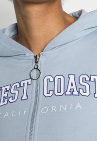 Sudadera con cremallera de color azul claro con capucha, que presenta texto en blanco y azul marino contrastante "WEST COAST" y "CALIFORNIA" en la parte frontal. Acento de cremallera plateada.