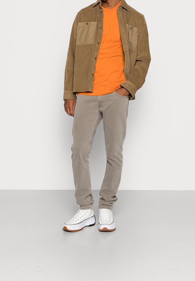 Homme portant une veste en velours côtelé beige sur une chemise orange, pantalon gris clair et baskets blanches avec des semelles marron, debout avec une main dans la poche.