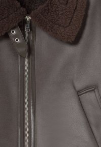 Veste en cuir marron avec un col en shearling marron, une fermeture éclair à l'avant et une poche latérale, le tout avec une texture lisse.