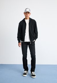 Čierna mikina s kapucňou na zips cez biele tričko, čierne slim-fit nohavice a čierne tenisky s bielymi akcentmi. Šedá čiapka dotvára celý outfit.