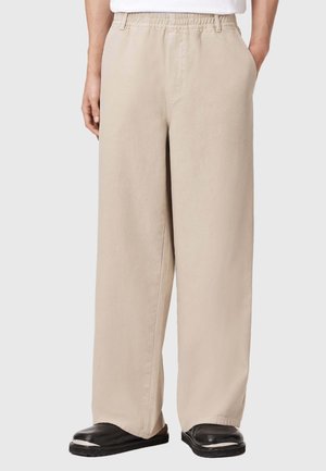 Pantalon beige à jambes larges avec ceinture élastique, porté par une personne debout, les mains dans les poches, associé à des chaussures noires sans lacets.