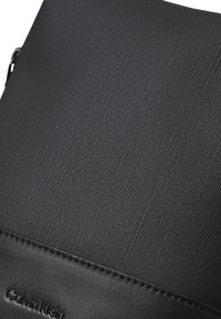 Bolso cruzado negro con un material superior texturizado, acentos de cuero suave y un logotipo sutil embellecido en el borde inferior.