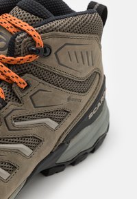 Brązowy but turystyczny z teksturowaną skórą i siateczkową górną częścią, z pomarańczowymi sznurowadłami, szarym wkładem i logo GORE-TEX. Ujęcie z bliska pod kątem.