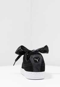 Černé semišové tenisky s texturovanou bílou gumovou podešví, vybavené saténovou černou mašlí na tkaničkách a stříbrným logem na patě.
