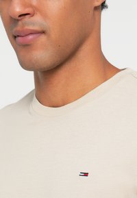 Μπέζ βαμβακερό t-shirt με στρογγυλή λαιμόκοψη, που διαθέτει ένα μικρό κεντημένο λογότυπο σε κόκκινο, άσπρο και μπλε χρώμα στο στήθος.