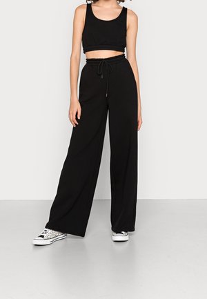 Frau trägt schwarzen ärmellosen Crop-Top, schwarze hoch taillierte weit geschnittene Hose mit Kordelzug und weiße flache Sneakers mit schwarzem Muster.
