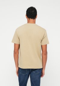 Beige puuvillane T-särk lühikeste varrukatega, ümar kaelus ja regulaarne lõige, nähtud tagant, valge tausta ees.