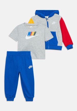 Παιδικό σετ τριών κομματιών Nike με γκρι μπλουζάκι με πολύχρωμο λογότυπο, μπλε παντελόνια φόρμας με λογότυπο και γκρι φούτερ με φερμουάρ, μπλε κουκούλα και κόκκινο μανίκι.