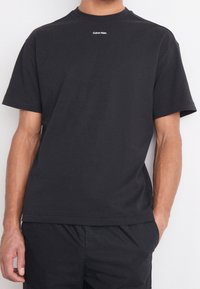 T-shirt en coton noir avec un col rond, des manches courtes et un petit logo blanc "Calvin Klein" sur la poitrine. Design simple.