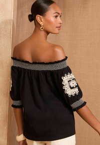 Top nero scollato con ricami floreali bianchi sulle maniche, colletto testurizzato e maniche a sbuffo. Materiale in cotone liscio.