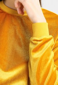 Sudadera de terciopelo amarillo con cuello y puños acanalados. La tela tiene una textura suave y brillante, reflejando la luz con un acabado liso.
