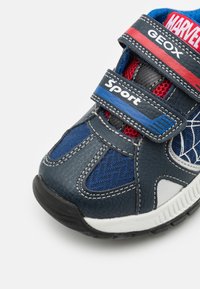 Blå och navy träningssko med läder och meshmaterial, kardborreband, röda accenter, vit sula och spindelnätsdesign på sidan.