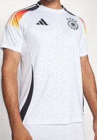 Biały strój sportowy z geometrycznym wzorem, czarnymi akcentami i kolorowym gradientem na ramionach. Zawiera logo adidas oraz herb Niemiec.