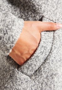 Tessuto di un cappotto grigio con una finitura caratterizzata da una superficie pelosa. Presenta una tasca laterale con una mano all'interno, che mette in mostra i materiali e i dettagli di design.