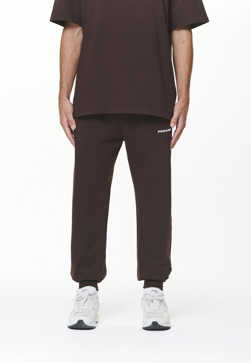 Camiseta de algodón marrón y pantalones deportivos a juego, con puños elásticos y un pequeño logo blanco en la pierna izquierda. Zapatillas blancas.