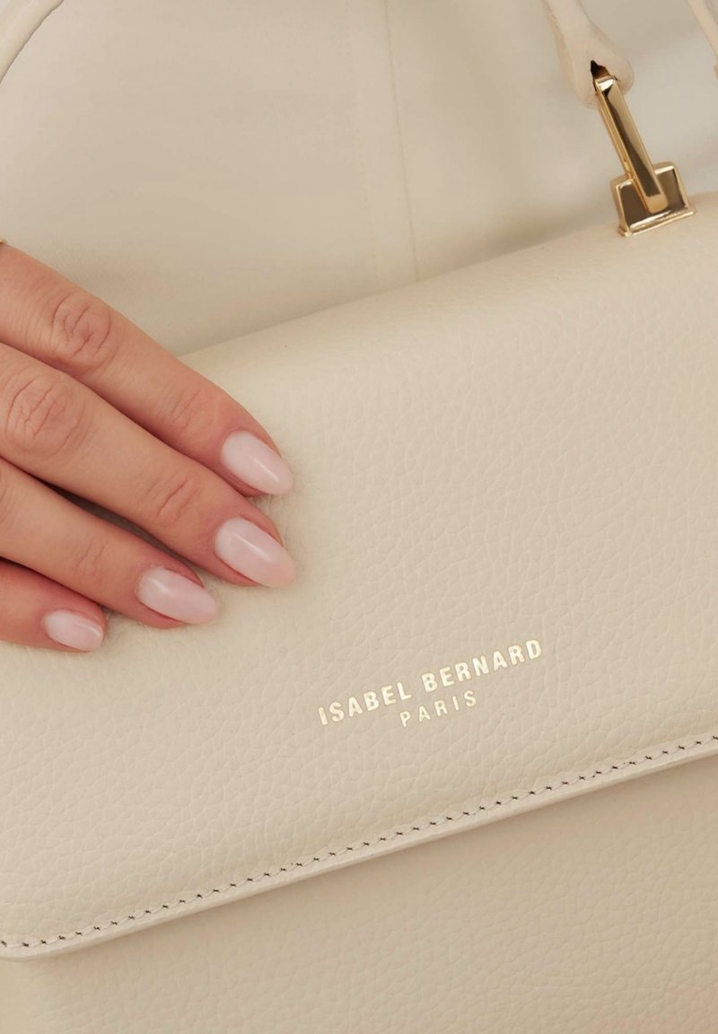 Main aux ongles manucurés rose pâle reposant sur un sac à main en cuir texturé crème avec un logo doré "Isabel Bernard Paris".