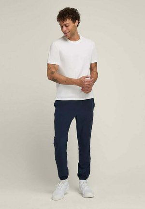 Weißes Kurzarm-T-Shirt, kombiniert mit navy Jogginghosen, die einen elastischen Bund haben, und weißen Sneakers. Das Model hat lockige Haare und sichtbare Tätowierungen.