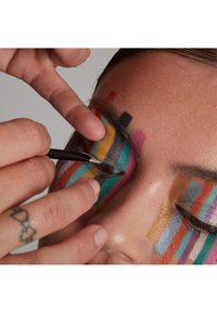 Heller, farbenfroher Gesichtsschminke mit mutigen Streifen und geometrischen Mustern. Eine Hand trägt mit einem feinen Pinsel Eyeliner auf und konzentriert sich auf das Auge.