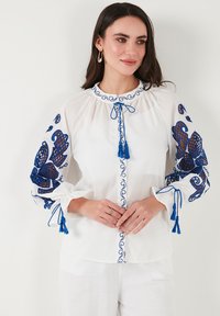Blusa bianca con farfalle ricamate blu e motivi. Caratterizzata da un collo rotondo, chiusura frontale con bottoni e dettagli con frange sulle maniche.