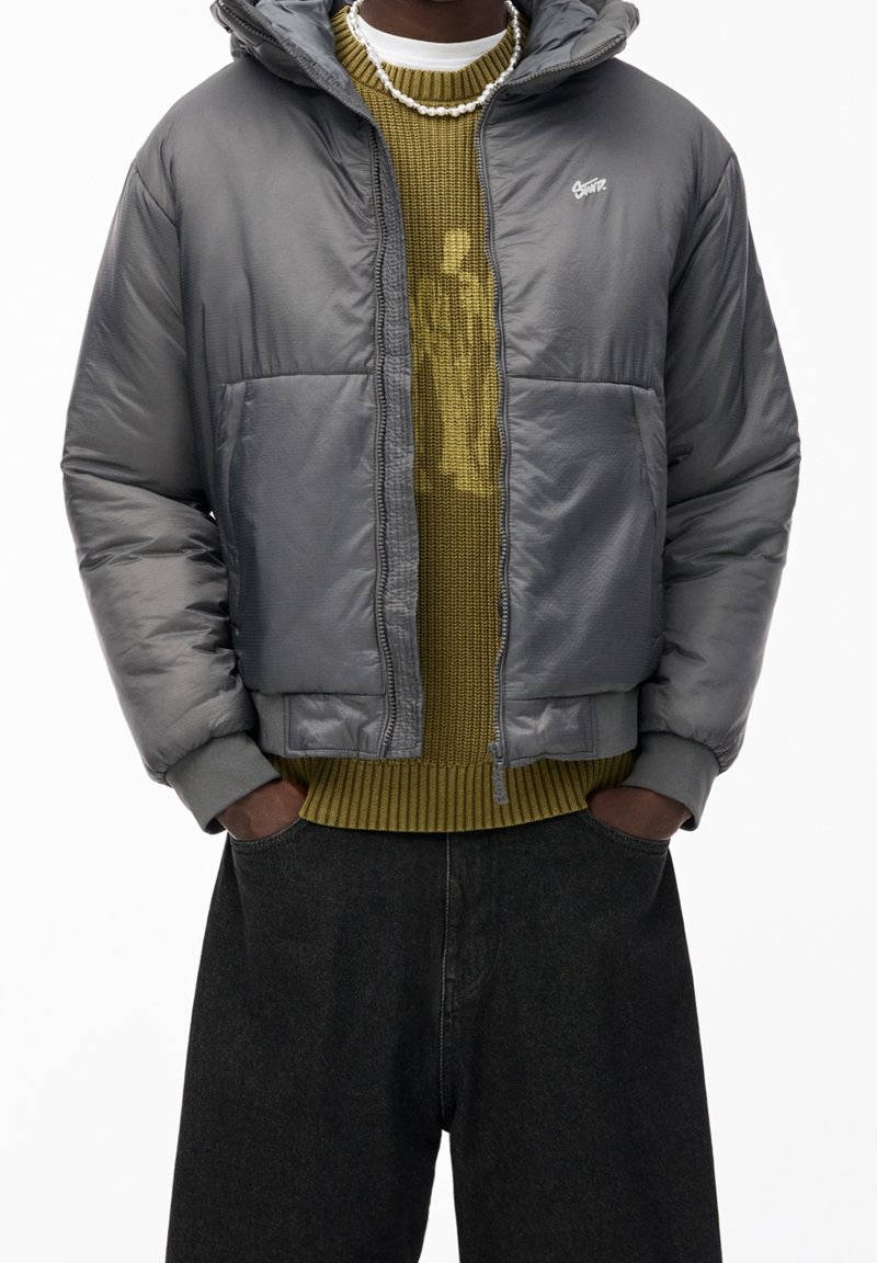 Veste grise isolante avec capuche, dotée d'une fermeture éclair et de deux poches. Portée sur un pull tricoté olive et un jean foncé ample.