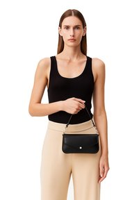 Borsa a tracolla nera in pelle con chiusura a patta e dettagli in argento, tenuta da una persona che indossa un top nero e pantaloni beige.