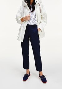 Chaqueta impermeable blanca con capucha, pantalones navy ajustados y zapatos azules con acentos rojos. Camiseta estampada en colores claros debajo de la chaqueta.