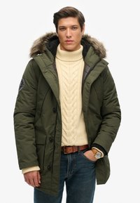 Parka acolchada verde aceituna con capucha de piel; presenta cierre de botones, bolsillos laterales y un suéter de punto texturizado en crema debajo.