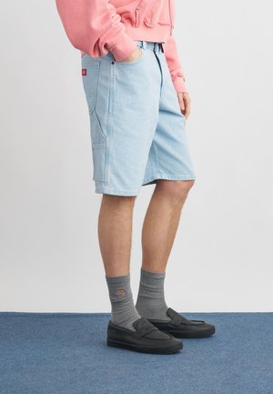 Hombre con pantalones cortos de mezclilla azul claro, sudadera rosa, calcetines grises con logo y zapatos negros sin cordones, de pie sobre una alfombra azul.