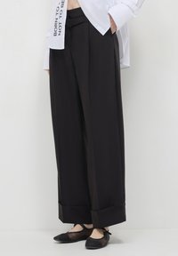Pantaloni neri a gamba larga con una superficie liscia, dotati di risvolto, passanti per cintura e una cintura abbinata per la chiusura. Abbinati a ballerine.