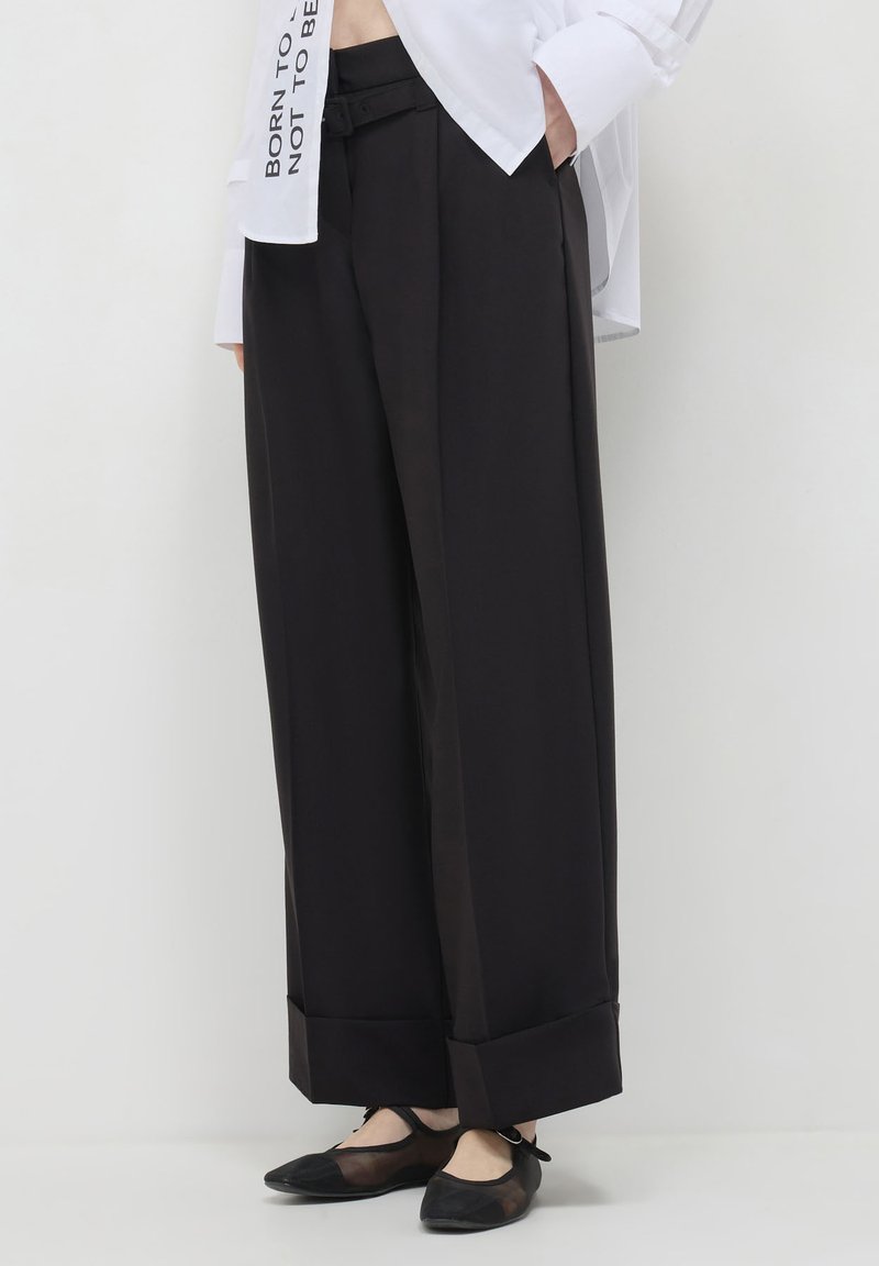 Pantaloni neri a gamba larga con una superficie liscia, dotati di risvolto, passanti per cintura e una cintura abbinata per la chiusura. Abbinati a ballerine.