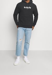 Sweat à capuche noir avec une poche avant et un logo "Levi's" blanc, assorti à un jean bleu clair déchiré et des baskets blanches.