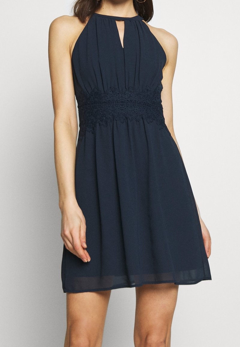 Femme portant une robe sans manches bleu marine à col licou, avec une ceinture en dentelle et un décolleté en trou de serrure, debout devant un fond clair uni.