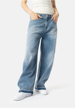 Person som har på seg lyseblå vide jeans, hvite sneakers og en hvit utenermet topp, med hendene i lommene.