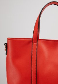 Sac à main en cuir rouge avec bords cousus, poignée verticale et petit anneau en métal doré sur le côté gauche sur fond uni.