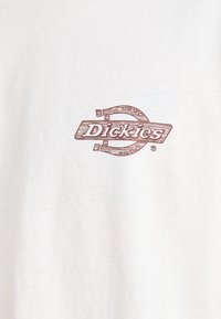 Bílá bavlněná tričko s hnědým grafickým logem se slovem "Dickies" uvnitř stylizovaného oválného tvaru. Hladká textura, klasický design.