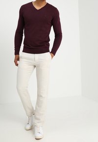 Burgunderfarbener V-Ausschnitt-Pullover, taillierte Passform. Kombiniert mit cremefarbenen Leinenhosen, gerader Beinabschluss. Weiße Sneakers mit beigen Akzenten.