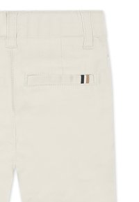 Pantaloni di cotone beige con una tasca posteriore caratterizzata da un'accente a strisce in nero, beige e bianco. Texture liscia e design su misura.