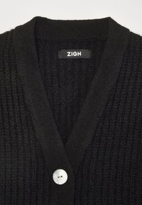 Černý pletený cardigan s výstřihem do V, který má výrazný texturovaný vzor a ozdobný bílý knoflík vpředu. Na štítku je napsáno "ZIGN."