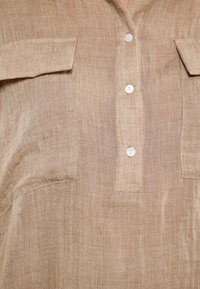 Camicia di lino marrone chiaro con chiusura a bottoni, due tasche sul petto e una vestibilità comoda, con texture e dettagli di cucitura visibili.