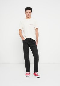 Λευκό βαμβακερό T-shirt με κόκκινο κείμενο, μαύρο τζιν παντελόνι και κόκκινα πάνινα παπούτσια. Το μοντέλο στέκεται ευθεία με τα χέρια στις τσέπες. Απλό φόντο.