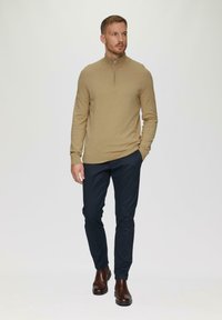 Un pull à fermeture éclair de couleur brun clair avec un col haut, associé à un pantalon slim en bleu foncé et des bottines brunes, se tenant contre un fond neutre.