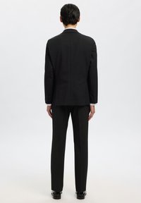 Czarny garnitur na miarę o smukłym kroju, z pojedynczym rozcięciem i minimalistycznym wykończeniem. Spodnie mają prosty krój, gładką fakturę i brak wzorów.