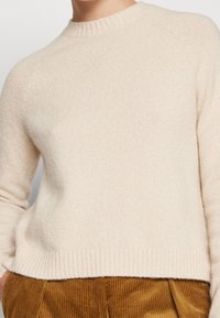 Maglione beige a maglia con colletto e fondo a costine, indossato con pantaloni in velluto a coste marroni, in cui si vedono il torso superiore e le mani nelle tasche.