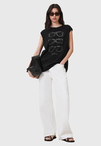 Schwarzes ärmelloses T-Shirt mit einem Grafikdesign von weißen Sonnenbrillen, kombiniert mit weißen weitem Hosen und einer schwarzen Tasche. Model trägt dunkle Sonnenbrillen.