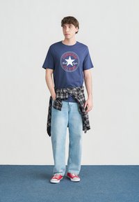 Jeune homme portant un t-shirt Converse All Star bleu marine, un jean bleu clair, des baskets rouges et une chemise à carreaux noire nouée autour de la taille.