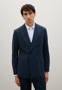 Blazer doppiopetto blu navy con rever affilati, due tasche frontali e una superficie liscia. Indossato su una camicia bianca con colletto.