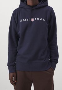 Persona con una sudadera con capucha azul marino que lleva "GANT 1949" y un logo de escudo en el pecho, combinada con pantalones marrones con texto sutilmente grabado.